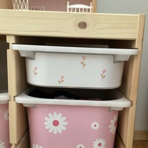 May include: A wooden shelf unit with two white and pink storage bins. The top bin is white with pink floral accents. The bottom bin is pink with white daisy patterns. The shelf unit is made of light-colored wood.