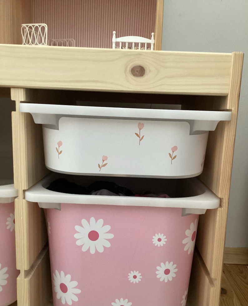 May include: A wooden shelf unit with two white and pink storage bins. The top bin is white with pink floral accents. The bottom bin is pink with white daisy patterns. The shelf unit is made of light-colored wood.