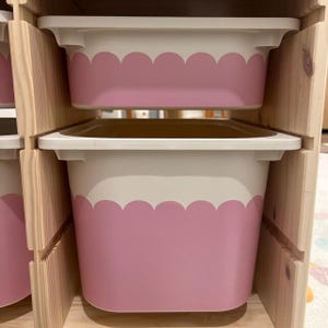 May include: Two white and pink storage bins with a scalloped design are stacked in a wooden shelf. The bins are rectangular with a white rim and a pink lower section. The wooden shelf has a natural finish.