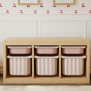 Könnte beinhalten: Ein helles Holzregal mit drei Fächern, in denen sich jeweils eine rosa-weiß gestreifte Box befindet. Die Boxen haben rosa Deckel. Der Hintergrund umfasst eine weiße Wand mit Kirschaufklebern und ein Bett mit gestreifter Bettwäsche.