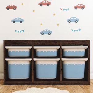 May include: A wooden storage unit with six cubbies, each holding a blue and white bin. The wall behind features car and cloud decals in red and blue, with yellow stars and bunting. A cloud-shaped rug sits on the wooden floor.