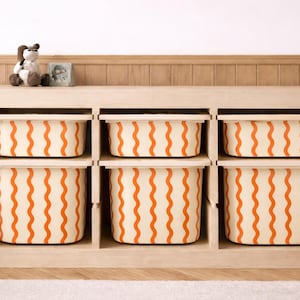May include: A light wood storage unit with six cream-colored bins. Each bin has a repeating orange wavy stripe pattern. A stuffed dog and small framed picture sit on top of the unit. The unit is on a light wood floor.
