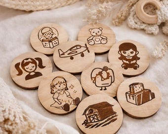 May include: A collection of nine wooden discs with engraved illustrations. The designs include a teddy bear, a girl reading, a plane, a girl, a girl with pigtails, a chicken, a barn, and a toaster. The discs are arranged on a white surface.