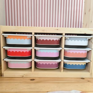 May include: A wooden storage unit with nine compartments, each holding a colorful bin. The bins have white rims and scalloped edges in various colors, including orange, red, pink, and blue. The background features a pink and white striped wall.