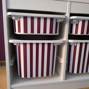 May include: A white storage unit featuring multiple open compartments. Each compartment holds a removable plastic bin with a vertical striped pattern in deep red and white. The bins are arranged in two columns, providing organized storage space.
