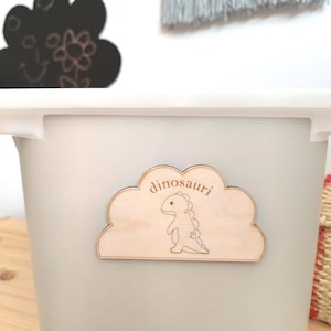 May include: A light grey storage bin with a white lid. A wooden cloud-shaped sign with the word "dinosauri" and a dinosaur illustration is attached. A black cloud-shaped chalkboard is visible in the background.