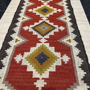 May include: A long, rectangular rug with a red central field and geometric patterns. The design includes diamond shapes in yellow, blue, and gray, bordered by a dark brown edge and white accents. The rug is likely made of woven material.