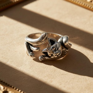 May include: A silver-colored ring featuring a stylized cat design. The ring wraps around the finger, with the cat's head and paws forming the focal point. The cat's tail curves around the band. The ring is set against a neutral background.
