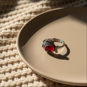 May include: A silver-toned adjustable ring featuring a fish design. The fish has a red, gem-like body and detailed fins and head. The ring is displayed on a beige plate, with a textured, off-white background.