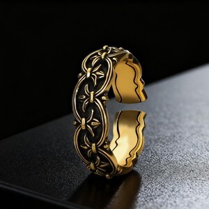 Puede incluir: Un anillo dorado con un diseño detallado. El anillo presenta un patrón oscuro y ornamentado en la banda y un diseño abierto único con siluetas de perfil. El anillo está sobre un fondo oscuro.