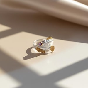 May include: A silver ring in the shape of a fish, featuring gold-colored fins and a purple gemstone eye. The ring is set against a neutral background, with shadows adding depth to the image. The fish design is detailed and artistic.