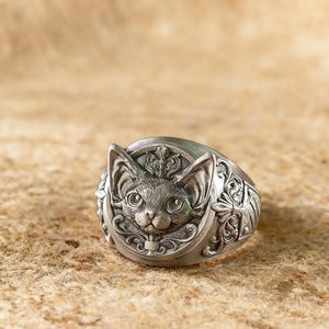 Puede incluir: Anillo plateado con un diseño detallado de cara de gato. El anillo presenta una cabeza de gato esculpida con orejas grandes y detalles intrincados alrededor de la cara y la banda. El anillo está sobre un fondo neutro.