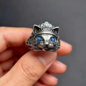 Op de afbeelding: Een zilverkleurige ring met een gedetailleerd kattengezicht, blauwe edelsteenogen en een kroon. De ring wordt in de hand gehouden tegen een donkere achtergrond, wat het ingewikkelde ontwerp en vakmanschap van de sieraden laat zien.
