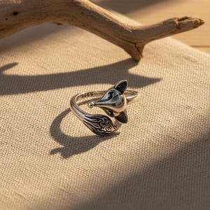Puede incluir: Anillo plateado con forma de zorro, con un diseño detallado de cabeza y cola. El anillo está sobre una tela de tono neutro, con una rama de madera en el fondo. La cola del zorro tiene un patrón intrincado.