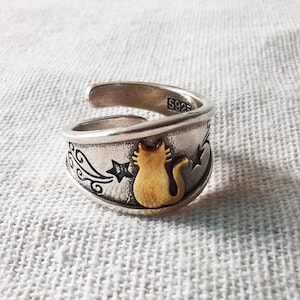 Puede incluir: Anillo ajustable plateado con silueta de gato dorada, estrellas y espirales decorativas. El anillo está estampado con "S925". El diseño del gato es el punto focal, sobre un fondo oxidado detallado. Una pieza de joyería única.