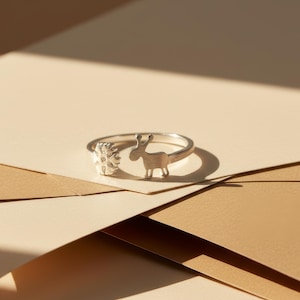 Peut inclure: Bague en argent avec un design fantaisiste. Elle présente un élément floral détaillé et une silhouette de cerf stylisée. La bague est mise en valeur sur un fond neutre, soulignant son artisanat délicat et son design.