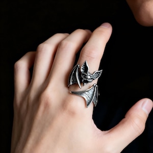 May include: A silver-colored bat ring with detailed wings and a bat face. The ring is wrapped around a finger, showcasing the design. The ring is a gothic-themed accessory.