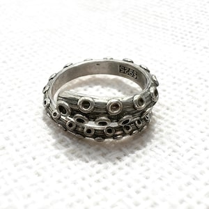 May include: A silver ring with an octopus tentacle design. The ring features a textured surface with multiple suction cup details. The band is open, and the ring is marked with "S925".