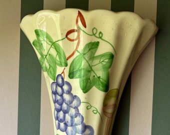 May include: A cream-colored ceramic wall pocket vase with a scalloped top edge. The surface features a hand-painted design of purple grape clusters, green vine leaves, and brown tendrils extending vertically down the center of the piece.