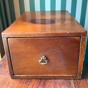 May include: A rectangular wooden storage box with a smooth, polished finish and a prominent dark wood grain knot on the top surface. The front features a single pull-out drawer equipped with a small, centered, antique-style brass drop handle.