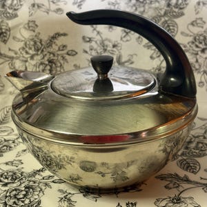 May include: A polished stainless steel kettle with a rounded body, a short spout, and a black curved handle. The lid features a small black knob at the center. The metallic surface reflects the surrounding light, showing a smooth, reflective finish.