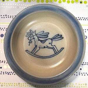 May include: A round ceramic bowl with a light beige interior and a dark blue rim. The center of the bowl features a dark blue line drawing of a rocking horse. The bowl has a smooth, glazed finish and a simple, circular shape with a wide edge.