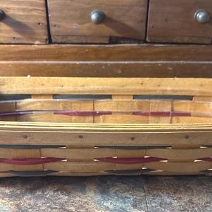 May include: Rectangular wooden basket with a clear plastic liner. The basket is woven with light brown wood and features red, black, and green accents. The basket is empty and sits on a stone surface.