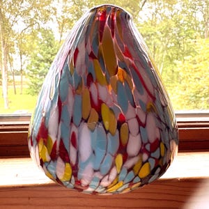 May include: A teardrop-shaped glass paperweight featuring a multi-colored confetti pattern with distinct flecks of red, light blue, yellow, and white embedded within the clear glass structure, creating a vibrant and speckled appearance.
