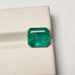 Könnte beinhalten: Ein loser Smaragd-Edelstein mit rechteckigem Smaragdschliff, der eine leuchtend grüne Farbe und facettierte Oberflächen aufweist, die das Licht reflektieren. Der Edelstein zeigt natürliche interne Einschlüsse und ein klares, poliertes Erscheinungsbild.