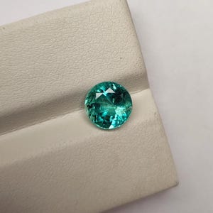 Könnte beinhalten: Ein runder, facettierter Edelstein in leuchtendem Türkisgrün. Der Stein ist im Brillantschliff gehalten und weist zahlreiche dreieckige Facetten auf, die das Licht reflektieren und für ein funkelndes Aussehen sorgen. Der Edelstein liegt auf einer strukturierten, cremeweißen Oberfläche.