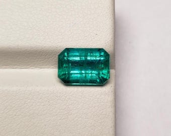 Puede incluir: Una piedra preciosa de corte esmeralda rectangular con un color verde vibrante e inclusiones internas visibles. La piedra presenta una superficie facetada que refleja la luz, mostrando una calidad clara y transparente con tonos verde intenso.