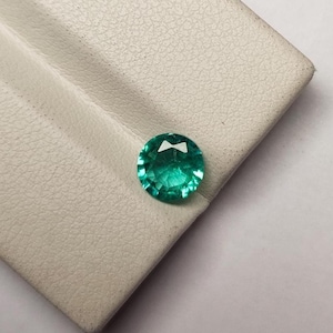 Könnte beinhalten: Ein runder, facettierter Edelstein in einer leuchtenden, durchscheinenden blaugrünen Farbe. Der Stein ist im Brillantschliff gehalten und weist zahlreiche dreieckige und rautenförmige Facetten auf, die das Licht reflektieren und funkeln.