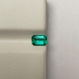 Op de afbeelding: Een rechthoekige smaragd met een levendige groene kleur. De edelsteen ligt op een getextureerd, gebroken wit oppervlak. De smaragd slijpvorm benadrukt de helderheid en schittering van de steen.