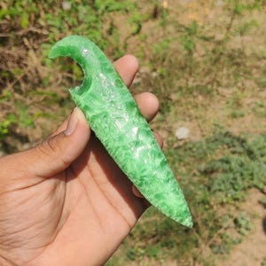 May include: A translucent green dagger with a textured surface. The object has a curved handle with a notch and a pointed tip. The dagger is held against a blurred background of green foliage.