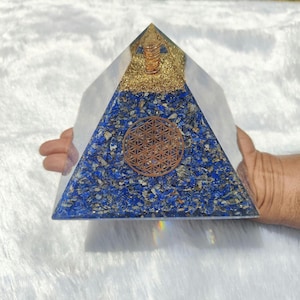 May include: A clear pyramid-shaped orgone device with a blue and gold design. The pyramid is filled with blue and gold stones and has a copper-colored geometric design on the front. The top of the pyramid is gold-colored.
