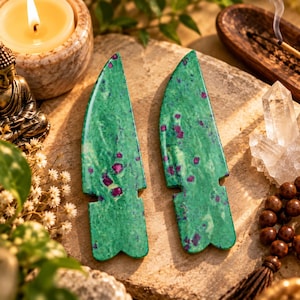 May include: Two polished green zoisite stone carvings shaped like stylized arrowheads or blades, featuring prominent magenta-colored ruby inclusions throughout the surface. The stones have a smooth, glossy finish and a distinct notched base design.