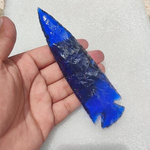 May include: A hand-held, blue glass arrowhead with a textured surface. The arrowhead is a deep, vibrant blue color, with a sharp point and a notched base. The object is likely a decorative or collectible item.