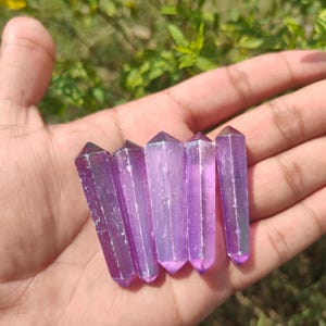 Puede incluir: Cinco varitas de cristal moradas con puntas afiladas se sostienen en una mano. Las varitas son de color morado translúcido y tienen un diseño facetado. El fondo es un follaje verde borroso.