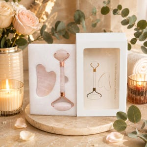 Puede incluir: Un set de masaje facial de cuarzo rosa que incluye un rodillo de doble extremo y una herramienta gua sha en forma de corazón en una caja blanca con ventana. La caja muestra un dibujo lineal de las herramientas y el texto Anti-aging Facial Massage Gift Set.