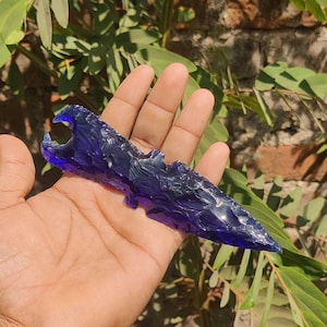 May include: A deep blue, hand-held, glass arrowhead with a textured surface. The arrowhead has a notched base and a sharp point. The object is held in a hand, with green foliage in the background.