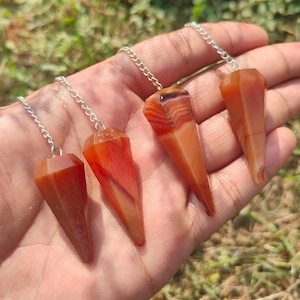 Op de afbeelding: Vier edelsteen slingerpendels met zilveren kettingen. De pendels hebben een mix van oranje en rode tinten, met facetvormige en puntige vormen. De achtergrond is een hand die de pendels vasthoudt, met groen gras zichtbaar.