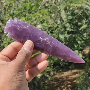 May include: A hand-held, purple arrowhead carved from stone. The arrowhead is a deep violet color with a pointed tip and a notched base. The background is blurred green foliage.