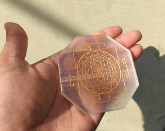 May include: A clear, octagonal selenite crystal with a gold-colored Sri Yantra design etched on its surface. The geometric pattern is centered within a square frame. The crystal is held in a hand, with a neutral background.