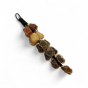 Puede incluir: Un adorno colgante decorativo con un grupo de piedras naturales en varios colores, incluyendo marrón, beige y verde. Las piedras están encerradas en una malla de alambre dorada y ensartadas en un cordón negro con un lazo en la parte superior.