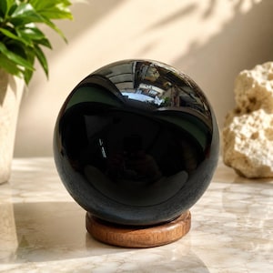 Op de afbeelding: Een gepolijste, bolvormige zwarte obsidiaansteen die op een ronde houten standaard rust. Het oppervlak van de bol is sterk reflecterend en toont vervormde reflecties van de omgeving, inclusief licht en architecturale elementen.