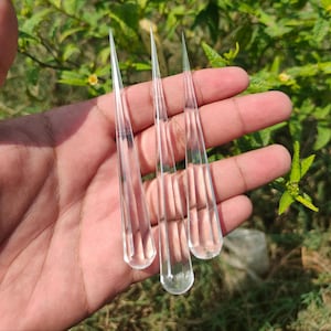Könnte beinhalten: Drei klare, tropfenförmige Glasornamente mit spitzen Spitzen. Die Ornamente werden in einer Hand vor einem Hintergrund aus grünem Laub gehalten. Jedes Ornament hat vertikale Linien, die in das Glas geätzt sind.