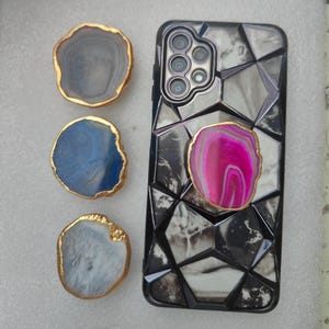 May include: A smartphone case with a geometric design and a pink agate stone phone grip. Three additional agate stones with gold edges are displayed. The agate stones are in shades of blue, gray, and white.