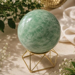 May include: A polished, spherical green stone with a marbled texture resting on a geometric gold-toned metal wire stand. The sphere displays varying shades of light and mint green with white veining throughout its smooth, reflective surface.