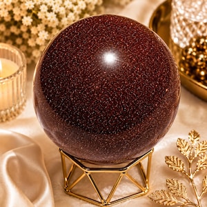 Peut inclure: Une grande boule de cristal rouge-brun étincelante repose sur un support géométrique doré. La sphère a un aspect scintillant. L'arrière-plan comprend une bougie, des fleurs et d'autres éléments décoratifs.