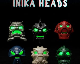 Può includere: Sei giocattoli Inika Heads in vari colori, tra cui grigio, nero, rosso, blu, bianco e verde. Ogni testa ha occhi verdi luminosi e un design unico. Il testo "VOYA-NUI SERIES" e "INIKA HEADS" è visualizzato in alto.